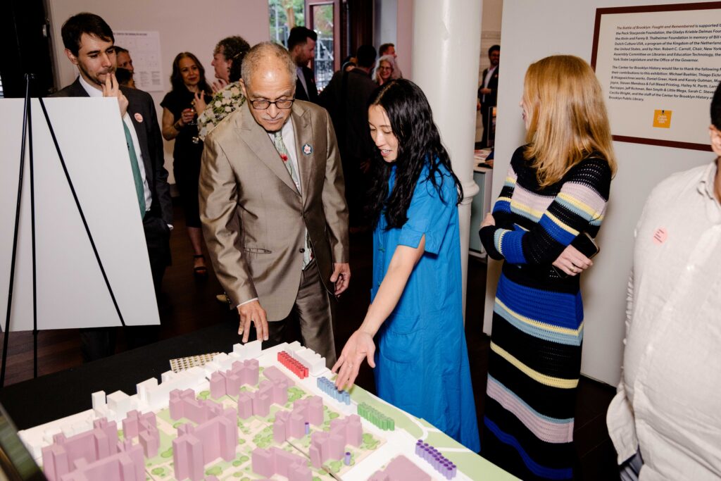 NY Chief Judge Rowan Wilson and Neighborhood Safety Initiatives Senior Director Layman Lee stand among a group of people looking at a miniature neighborhood model on display.