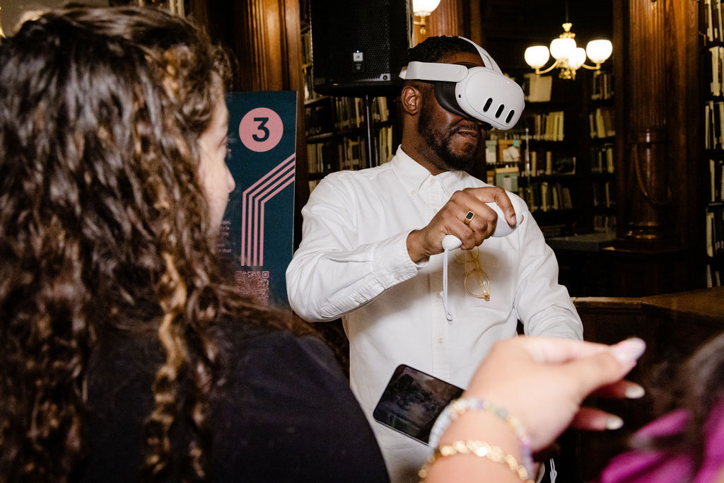 A man is shown wearing a VR headset and remote in an indoor space as a woman looks on while holding a smartphone.