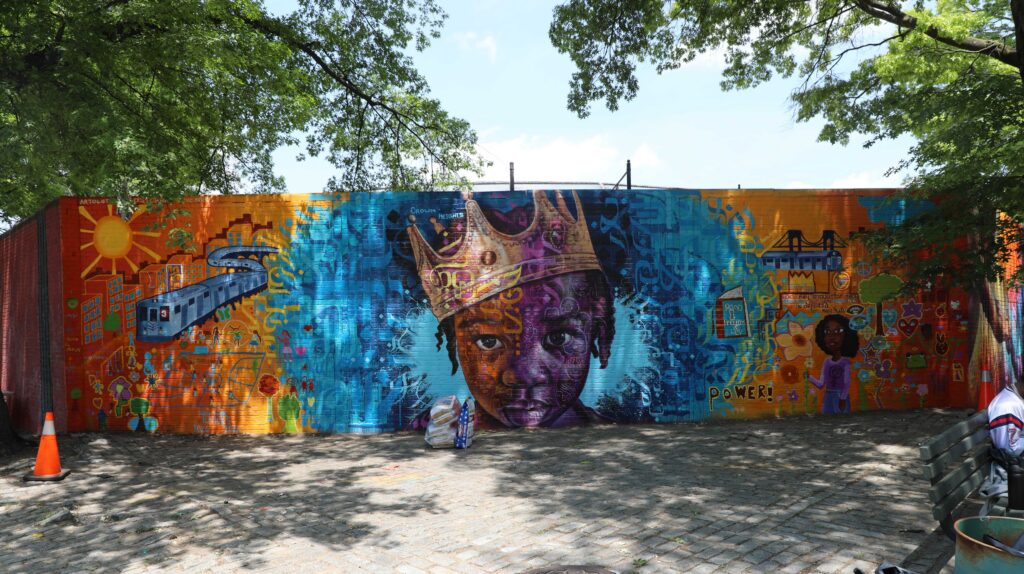 Wide-shot of a mural across a brick wall featuring a painting of a young Black boy wearing a crown looking outward; on the sides are depictions of various sights including a train, bridge, children playing, a young Black woman holding a flower, and apartment buildings.