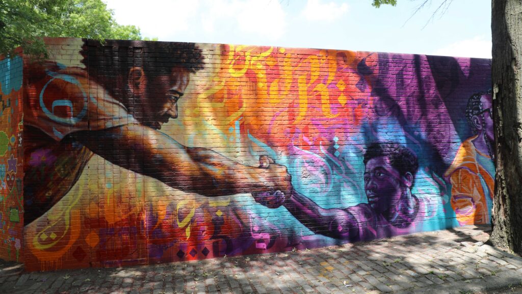 Mural on brick wall depicting a man stretching out his arm and grasping the hand of another man, situated lower; in the backdrop, decorative lines and font float over patches of orange, purple, and blue.