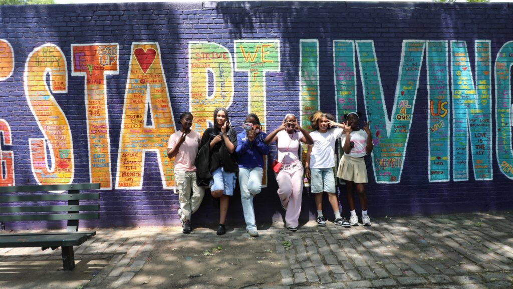 A group of young people pose making peace signs in front of a colorful mural with the words "Start Living" in bold letters