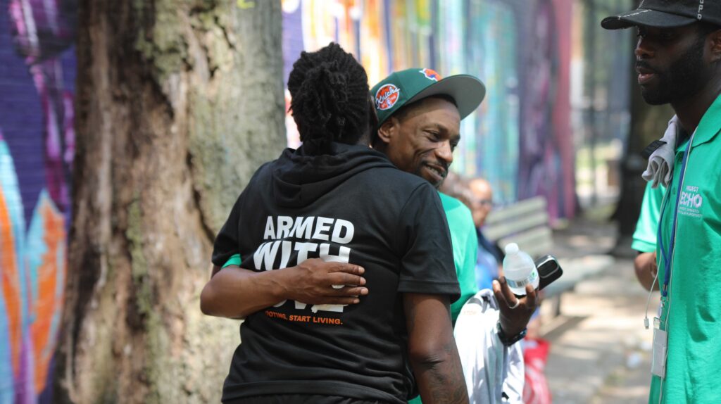 Two people hugging outdoors; one wears a black Save Our Streets sweatshirt, the other a green T-shirt with a New York Knicks hat.