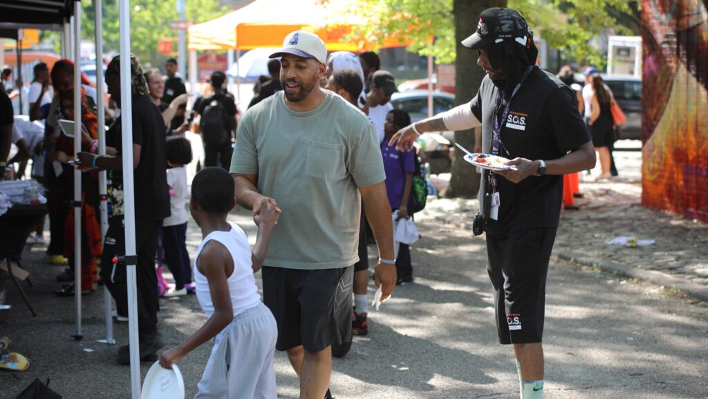 A father holds the hand of his young son, who is carrying a frisbee; behind, people line up at an outdoor resource fair