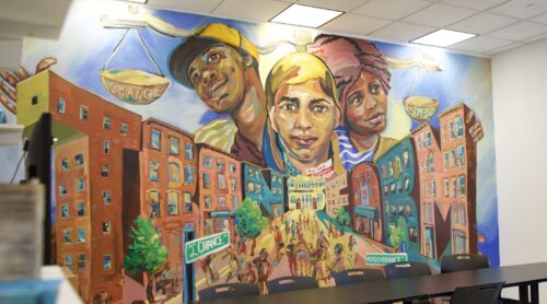 A mural at the Red Hook Community Justice Center features three people peering out from beyond a lively city street with a street sign that says 