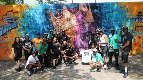 A group of women and men with our Save Our Streets team pose for a group picture in front of a mural featuring a Black child wearing a crown; in the middle, one man holds up a T-shirt that reads 
