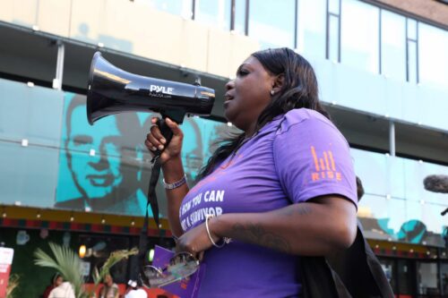 Woman in a purple RISE t-shirt speaks into a megaphone.
