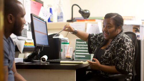 A woman sits in an office in conversation with a man seated beside her desk; on top of the desk is a computer and a sign that says 