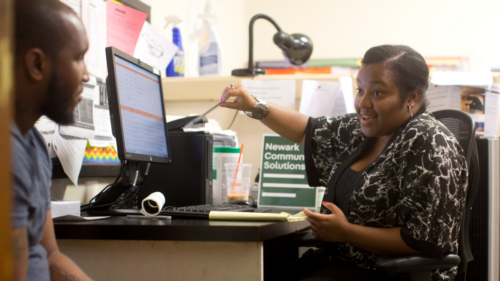 A woman sits in an office in conversation with a man seated beside her desk; on top of the desk is a computer and a sign that says 