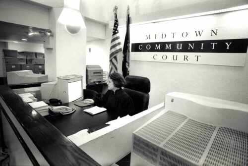 Black and white image of judge sitting on the stand on the computer with Midtown Community Court in the background