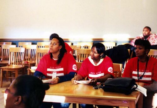 Group of e women sitting at a table in the Red Hook Community Justice Center court room with red shirts on.